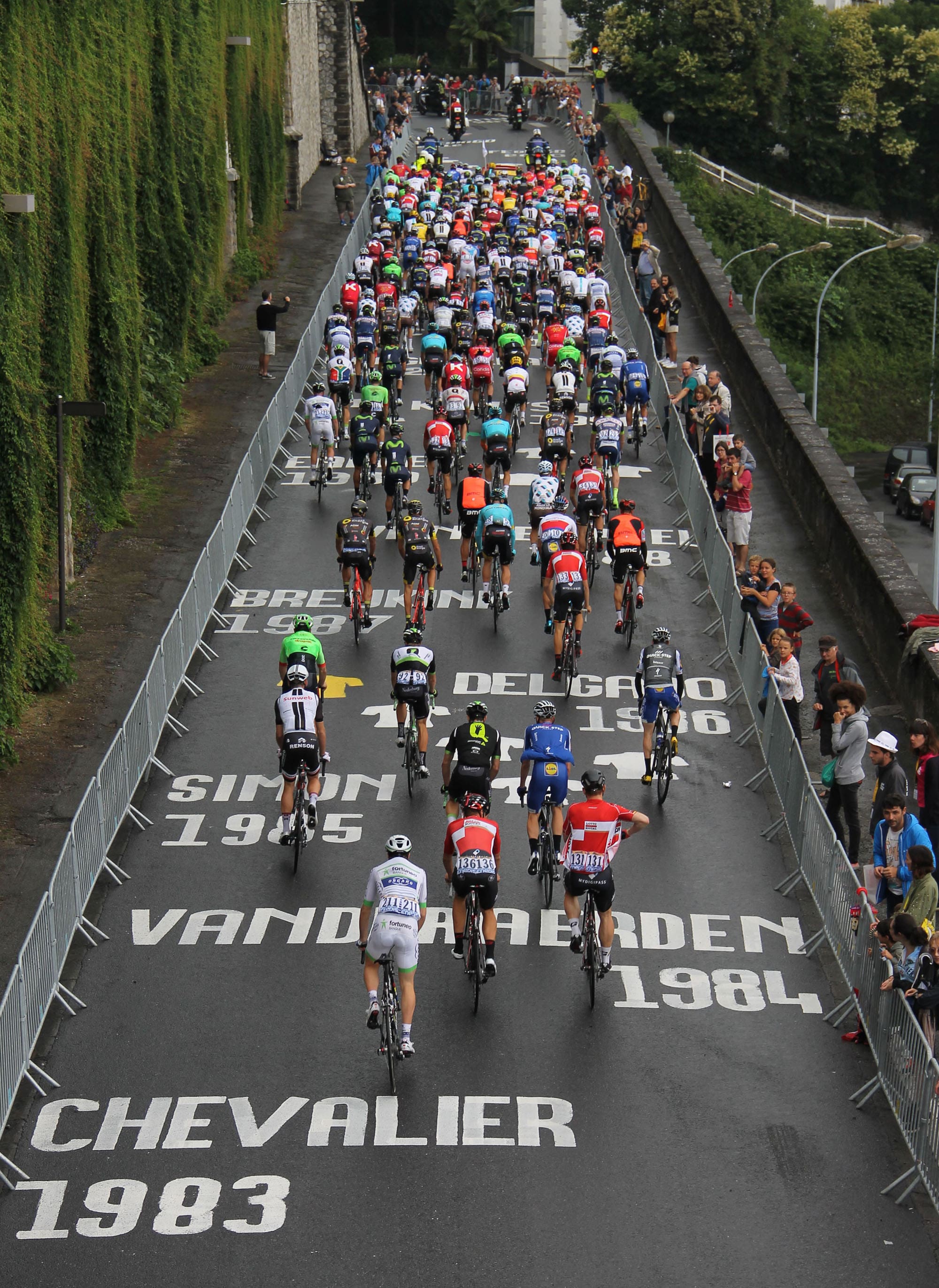 Photographie d'une cinquantaine de coureur cycliste remontant une route sur laquelle est peinte plusieurs noms et dates de gagnant d'étapes du Tour de France